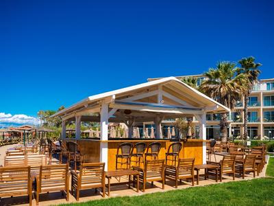 Outdoor bar with wooden furniture, palm trees, and hotel in the background under clear sky.