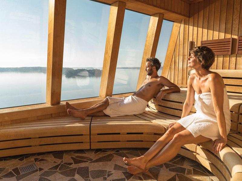 Two people relax in a sauna with large windows overlooking a lake.