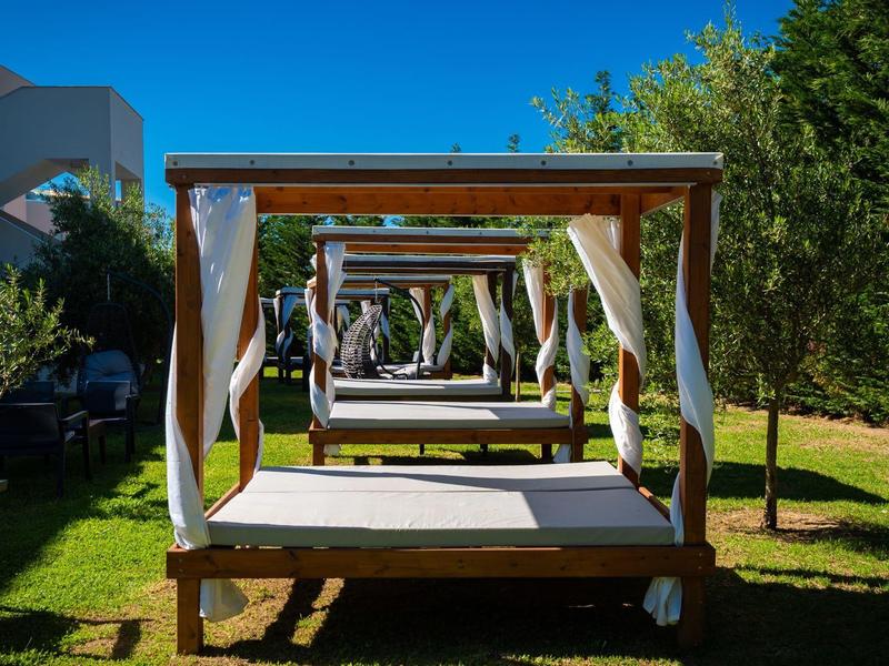 Rows of wooden lounge beds with white drapes on grassy area under blue sky.