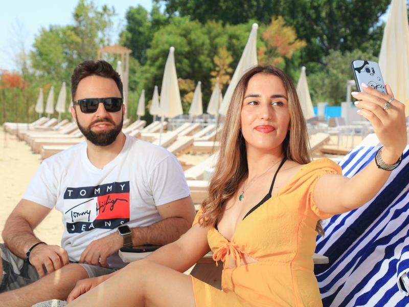 Due persone in spiaggia con lettini e ombrelloni, una si fa un selfie.