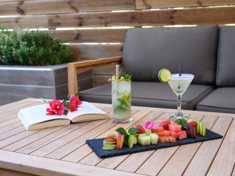 Holztisch mit frischem Obst, zwei Cocktails, geöffnetem Buch und Sofa im Hintergrund auf Terrasse.