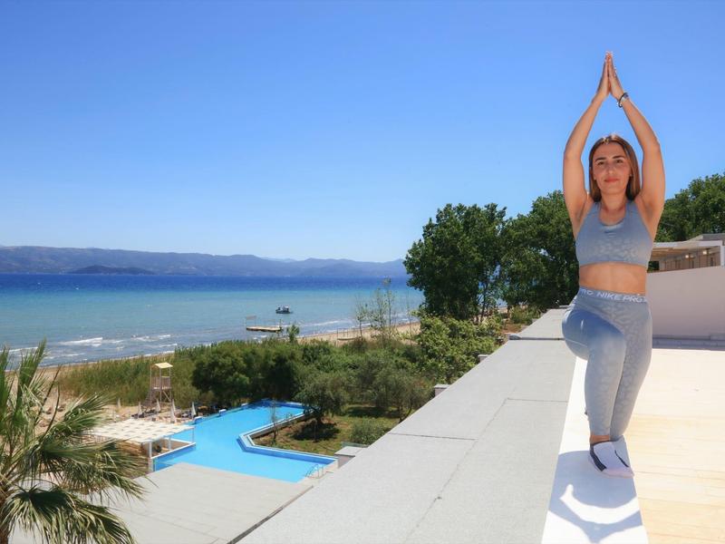 Frau macht Yoga auf Dach, blauer Himmel, Meer, Pool und grüne Pflanzen im Hintergrund.