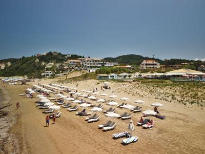 Sandstrand mit Reihen von weißen Sonnenschirmen und Liegen vor einem Hotel am Hügel.