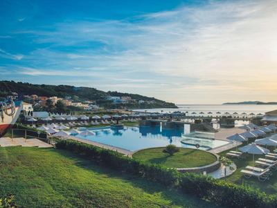 Panorama di un hotel con piscina e lettini accanto a una marina al tramonto.