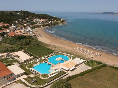 Küste mit Sandstrand, Poolanlage, Sonnenschirmen und Häusern an einem bewaldeten Hügel.