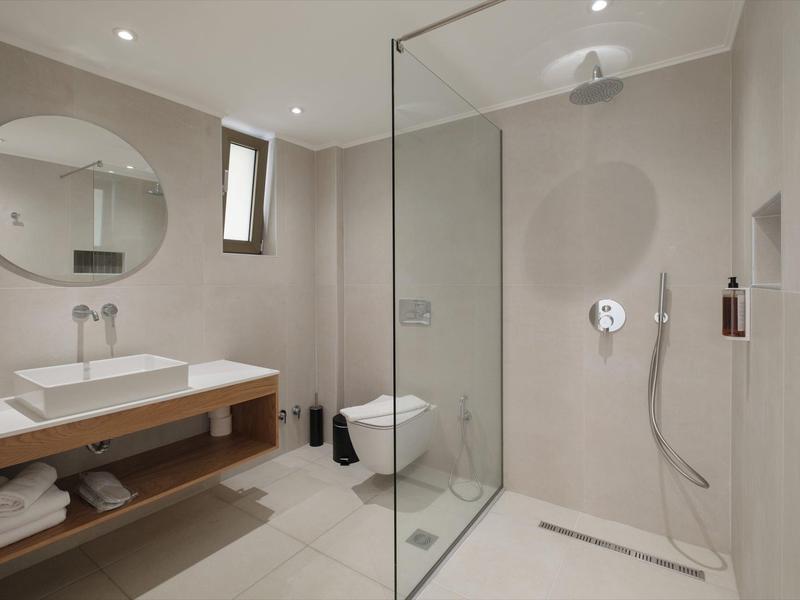 Modernes Badezimmer mit beiger Fliesenoptik, großer Glasdusche und rundem Wandspiegel.