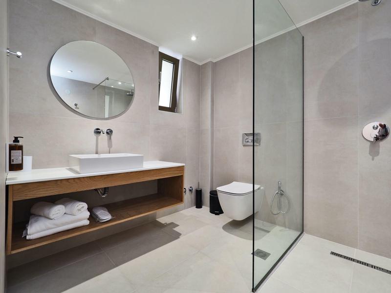 Modernes Badezimmer mit hellen Fliesen, Glasdusche, Wand-WC und Holzregal unter dem Waschbecken.