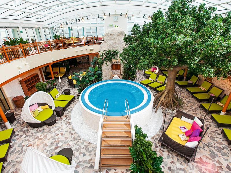 Heller Innenbereich mit kleinem Pool, großen Bäumen, bunten Sitzmöbeln und einem gläsernen Dach.