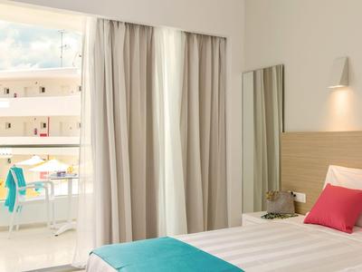 Camera d'hotel luminosa con letto bianco, copriletto turchese e balcone con vista sul mare.