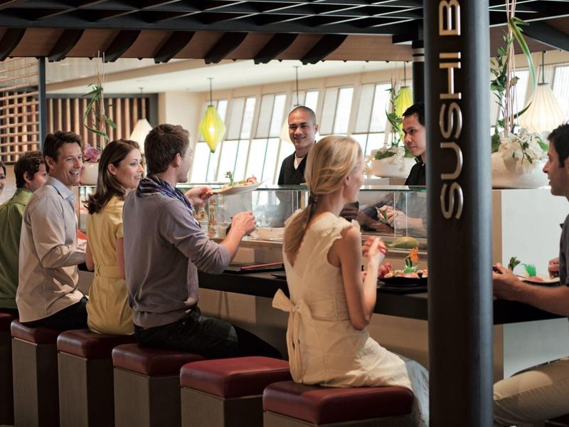 Menschen sitzen an einer Sushi-Bar, unterhalten sich und essen, helle und warme Innenraumbeleuchtung.