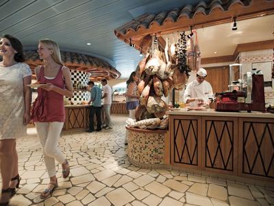 Two women walk in a hotel market area with a butcher stand and Mediterranean decor.