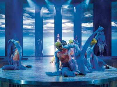 Dancers with colorful body paint on a round stage against a sky backdrop.
