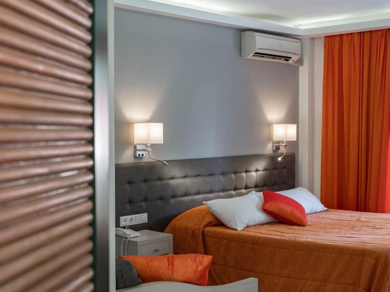 Hell eingerichtetes Schlafzimmer mit orangen Vorhängen und Bettdecke, grauem Kopfteil und Wandlampen.