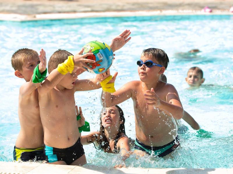 Kinder spielen im Schwimmbecken mit Wasserbällen, tragen bunte Armbänder und schwimmen im blauen Wasser.