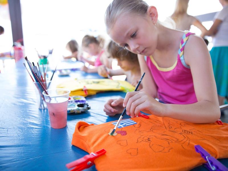 Kinder malen konzentriert auf orangefarbene Stofftaschen an einem hellen Basteltisch.