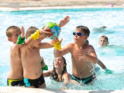 Kinder spielen im Schwimmbecken mit Wasserbällen, tragen bunte Armbänder und schwimmen im blauen Wasser.