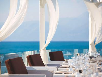 Elegante Terrasse am Meer mit weißen Vorhängen, braunen Sesseln und gedeckten Tischen.