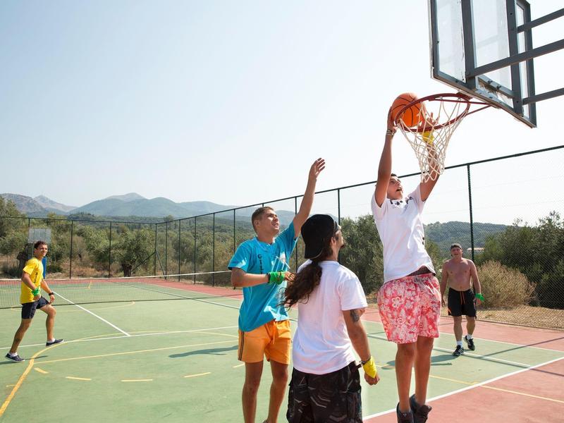 Kinder spielen Basketball auf einem sonnigen Außenplatz mit Bergen im Hintergrund.