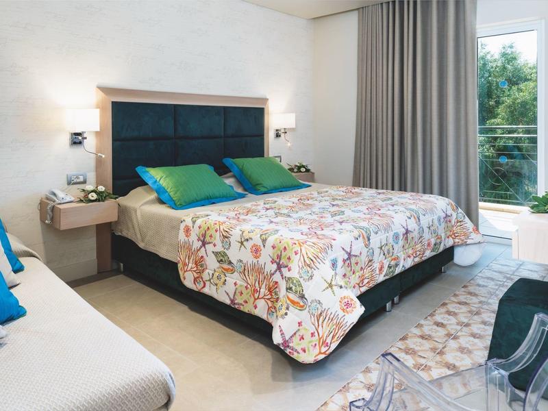 Hell eingerichtetes Schlafzimmer mit großem Bett, buntem Bettbezug, grauen Vorhängen und großem Fenster.