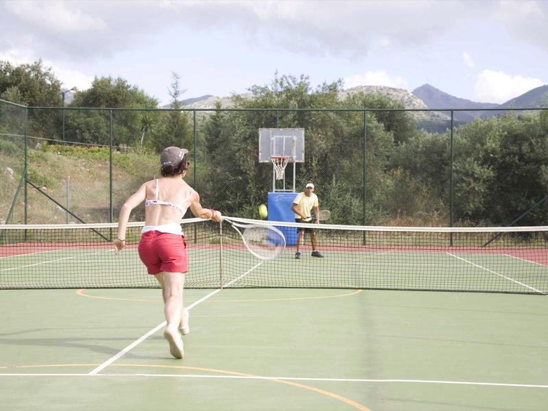 Zwei Jungen spielen Tennis auf einem Außenplatz mit grünem Boden und Zaun im Hintergrund.