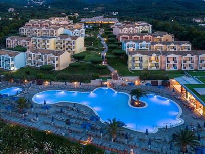 Große Hotelanlage bei Nacht mit beleuchtetem Pool, umgeben von mehrstöckigen Gebäuden und grüner Landschaft.