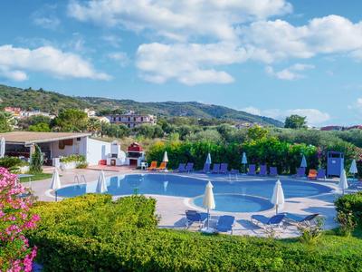Poolbereich mit Sonnenschirmen, Liegestühlen und grüner Umgebung unter blauem Himmel.
