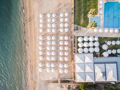 Vista aerea di una spiaggia con file di ombrelloni bianchi accanto a piscina e edifici.