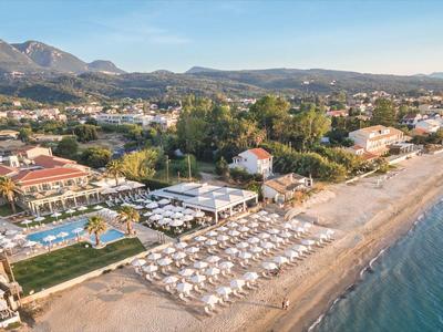 Strand mit Liegestühlen und Sonnenschirmen neben einem Resort am Wasser, Berge im Hintergrund.