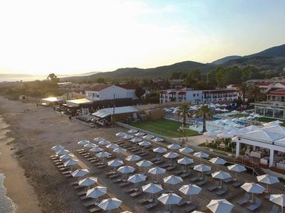 Strand mit Sonnenliegen und Schirmen neben einem Hotelkomplex in hügeliger Umgebung bei Sonnenuntergang.