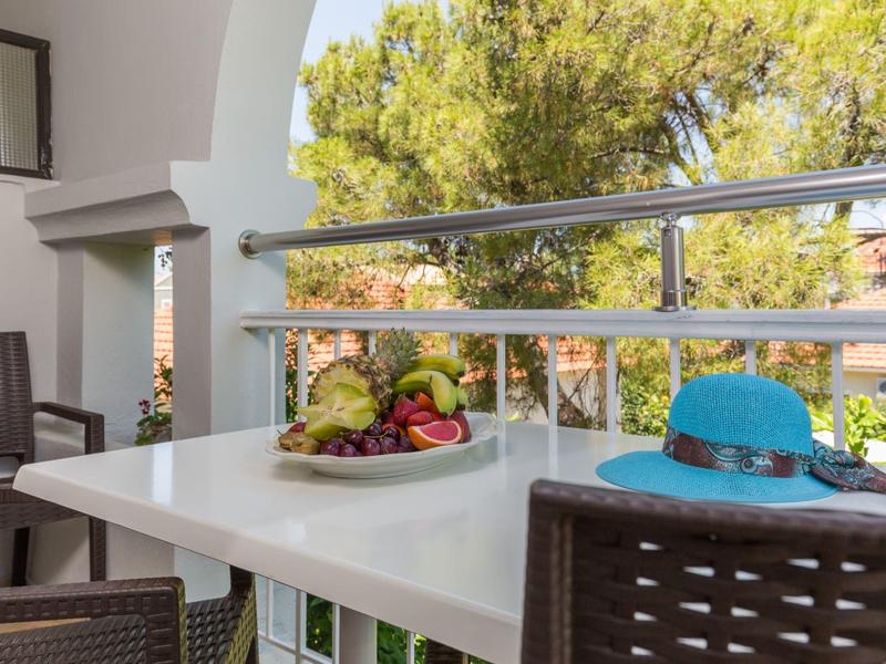 Ein gedeckter Balkon mit Tisch, Stühlen, Obstteller und blauer Sonnenhut neben grünen Bäumen.