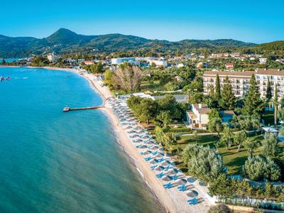 Wybrzeże z piaszczystą plażą, parasolami i hotelami na tle pagórkowatego krajobrazu pod klarownym niebem.