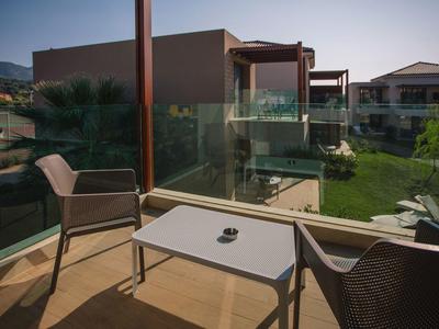 Balkon mit Glasgeländer, zwei Stühlen und Tisch, grüne Wiese und Häuser im Hintergrund.