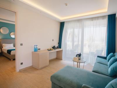 Helles Hotelzimmer mit blauem Sofa, Schreibtisch, Bett und großen Fenstern mit blauen Vorhängen.