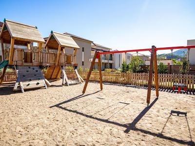 Spielplatz mit Sandsboden, zwei Holz-Schaukelgerüsten, Kletterwand und mehreren Spielhäusern.