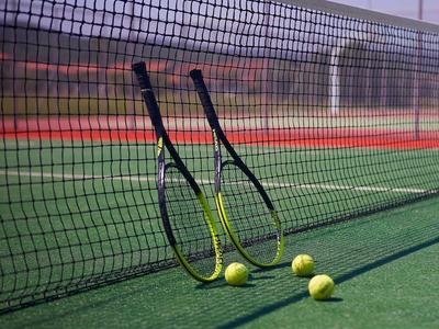 Zwei Tennisschläger lehnen am Netz eines Tennisplatzes mit drei Tennisbällen auf grünem Boden.