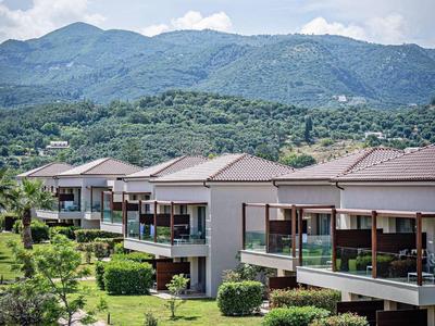 Moderne Häuser mit großen Balkonen in grüner Landschaft vor bewaldeten Bergen und blauem Himmel.
