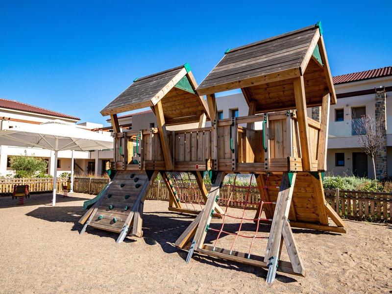Kinderspielplatz mit zwei Holzhäuschen, Kletterwänden und Seilrutschen vor blauem Himmel.
