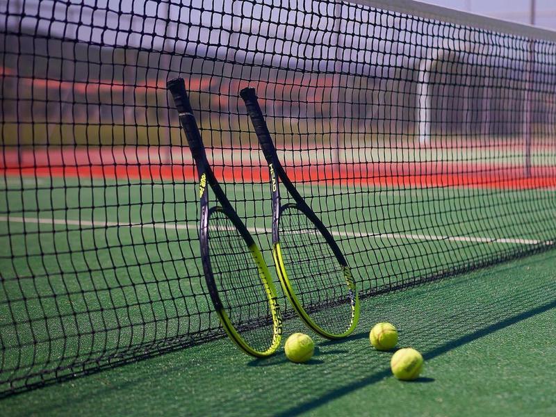 Tennisnetz mit zwei Schlägern und drei Tennisbällen auf grünem Platz bei klarem Himmel.