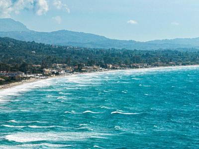 Weite Küstenlinie mit sandigem Strand, blauem Meer und bewaldeten Hügeln im Hintergrund.