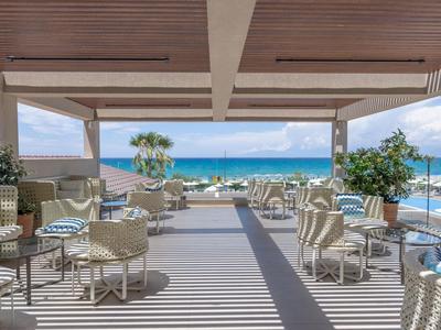 Offene Terrasse mit weißen Korbmöbeln, Pflanzen und Blick auf das blaue Meer unter blauem Himmel.