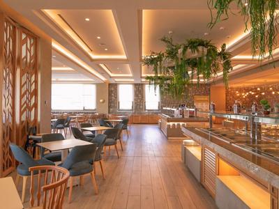 Modernes Restaurant mit Holzboden, grünen Pflanzen und gemütlichen Sitzbereichen in warmem Licht.