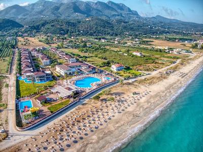 Landschaft mit Hotelanlage, Pool, Strand mit Sonnenschirmen und Bergen im Hintergrund unter blauem Himmel.