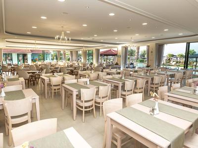 Großer heller Speisesaal mit vielen weißen Holztischen und Stühlen, großen Fenstern und grünen Tischläufern.