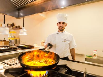 Koch in weißer Uniform steht an Herd, Pfanne mit Flammen, moderne helle Küche.