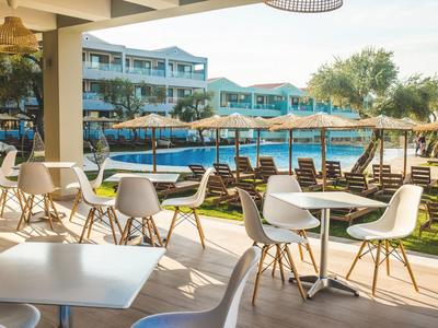 Freiluftrestaurant mit weißen Tischen, Stühlen und Blick auf einen Pool mit Liegen und blauen Sonnenschirmen.