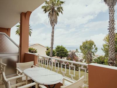 Balkon mit weißem Tisch und Stühlen, Palmen und Meer im Hintergrund bei bewölktem Himmel.