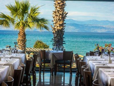 Restaurant mit gedeckten Tischen, Palmen und Blick auf das blaue Meer und Berge im Hintergrund.