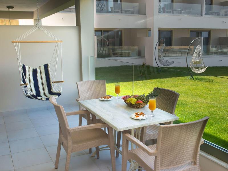 Moderner Balkon mit weißem Tisch, vier Stühlen, Hängesessel und gläserner Brüstung, Blick auf Grünfläche.