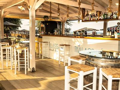 Hell beleuchtete Bar mit weißen Holzmöbeln und Strohsitzen unter offenem Dach.