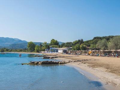 Strand mit klarem blauem Wasser, Sand, Sonnenschirmen und Bäumen im Hintergrund unter blauem Himmel.